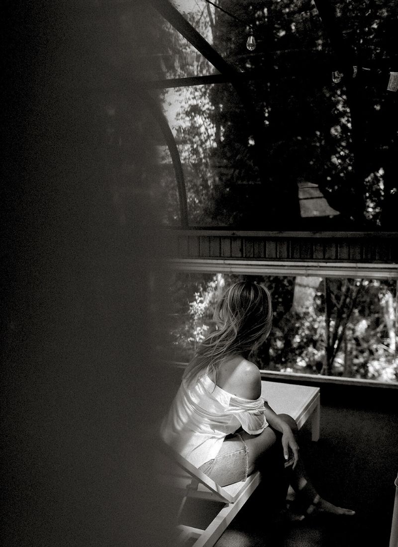 Woman sitting in a serene glass greenhouse, gazing into nature, embodying quiet feminine energy and self-reflection.