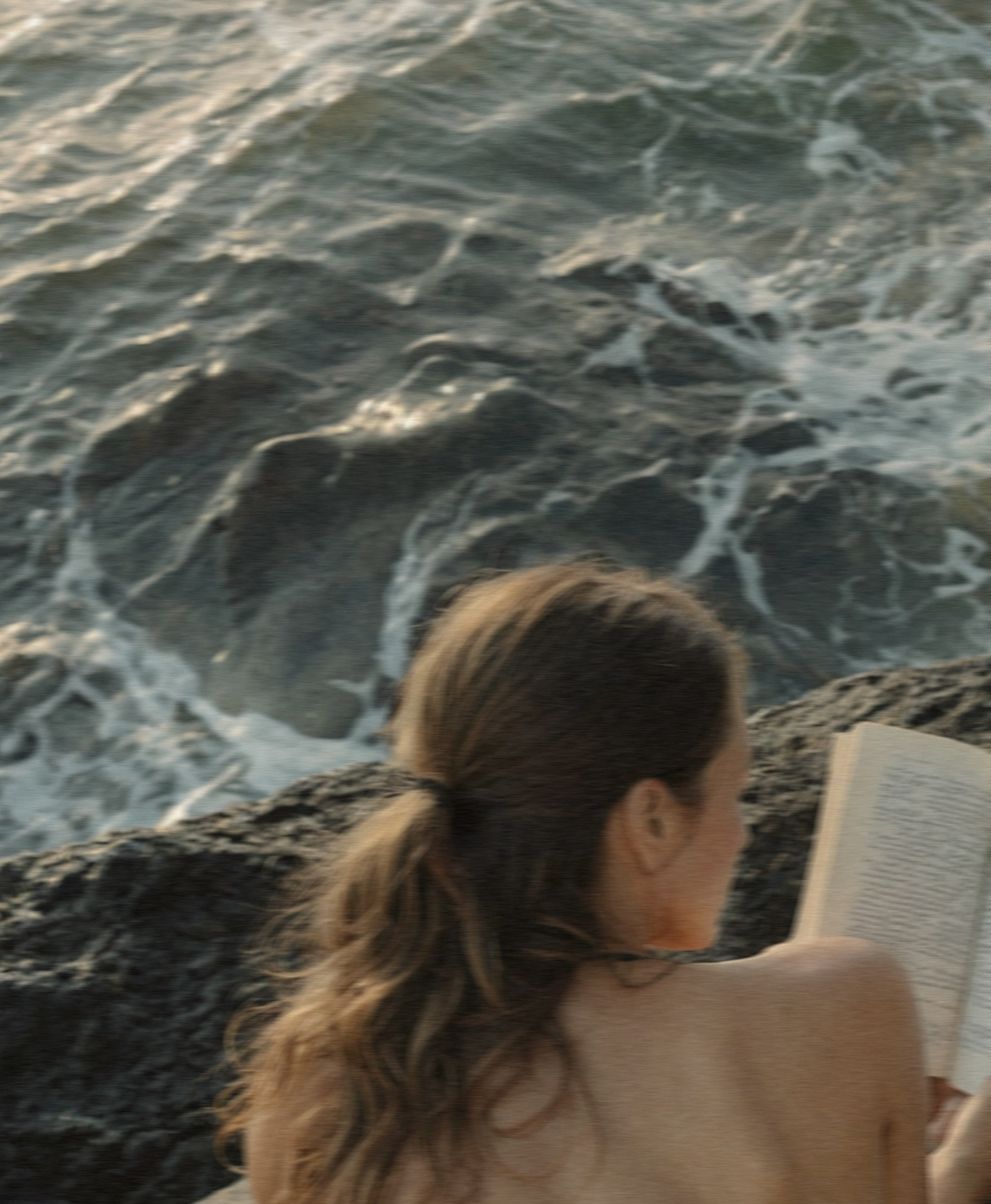 Woman reading a book on a rocky coast with ocean waves in the background, evoking wanderlust and introspection