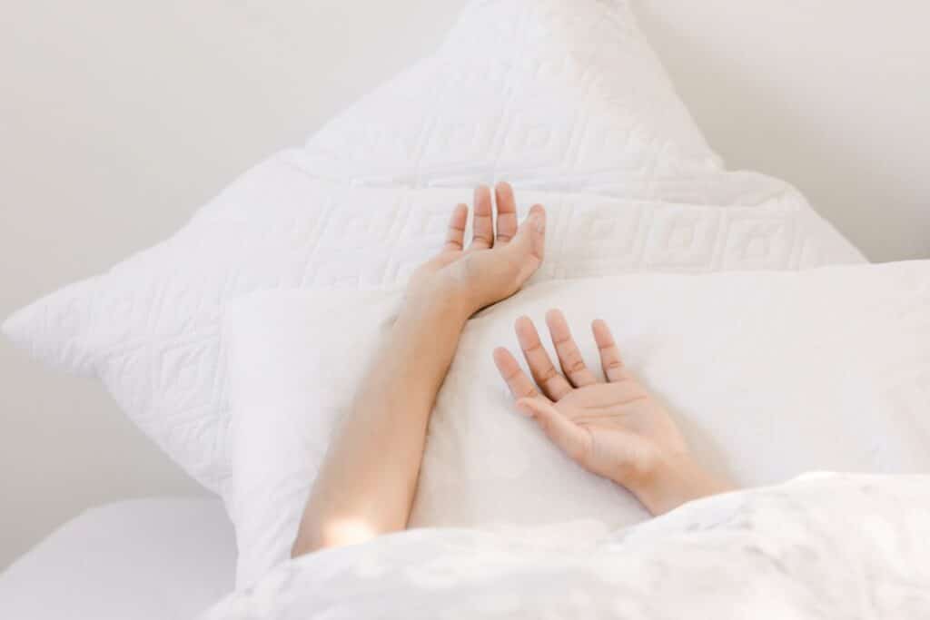 A person's hands gently rest on a white pillow, partially covered by a soft duvet, symbolizing peaceful sleep and the essential comfort of a well-prepared bed, embodying the essence of beauty sleep essentials.