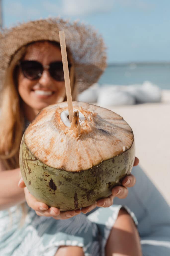 A smiling woman in a sun hat offers a fresh coconut with a straw on a sandy beach, epitomizing the tropical luxury of Mauritius 5 star hotels.