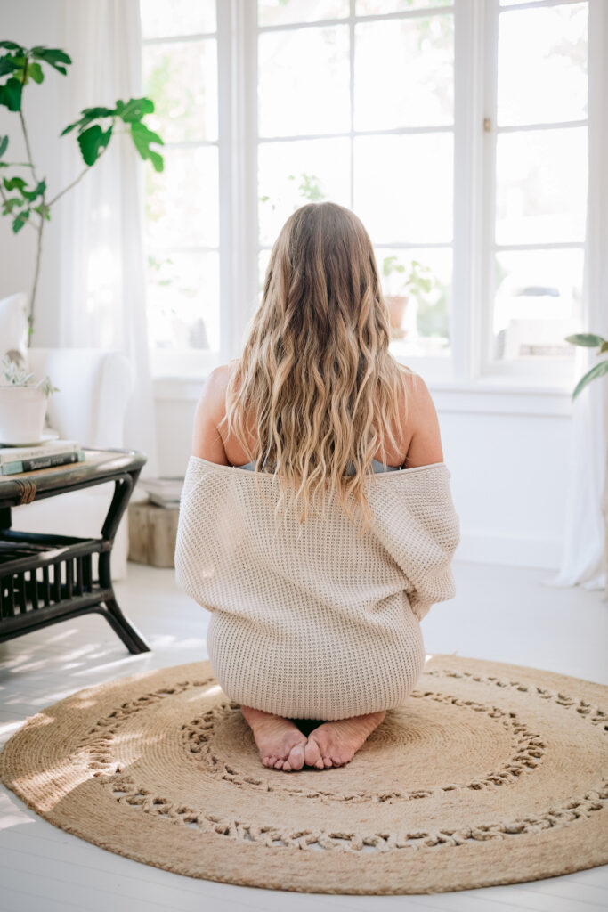 A woman sits peacefully in a sunlit room, draped in a cozy sweater, embodying tranquility and self-reflection, which are key to recognizing signs of blocked feminine energy.