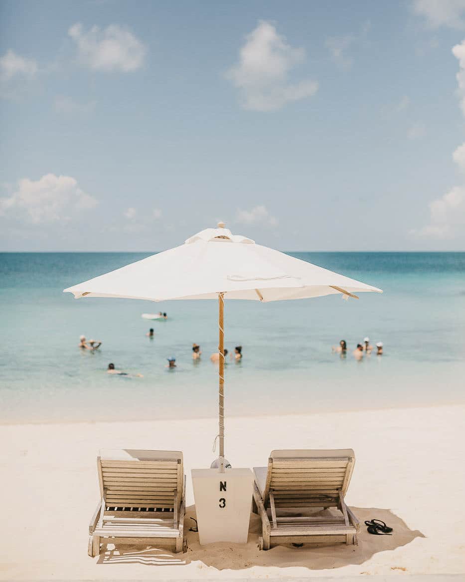 wo lounge chairs under a white umbrella on a pristine beach, with the ocean and swimmers in the background, ideal for captions about Vitamin Sea Quotes for Instagram.