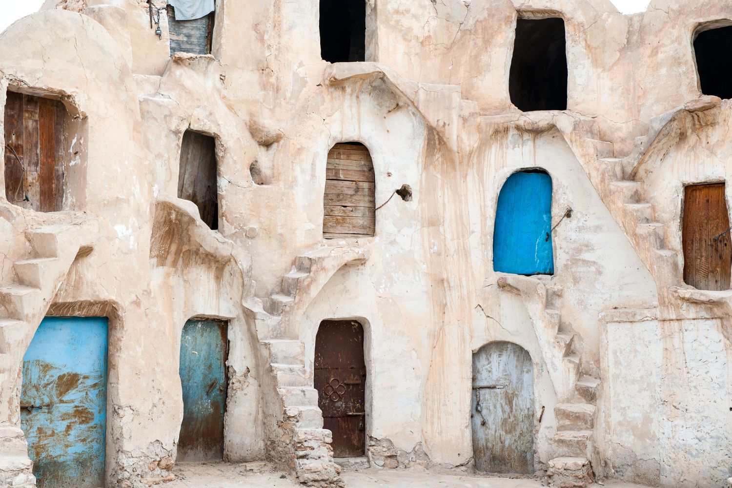Is Djerba worth visiting? Ancient troglodyte dwellings with textured clay walls and varying door shapes, some painted in bright blue, in the traditional architectural style of Djerba.
