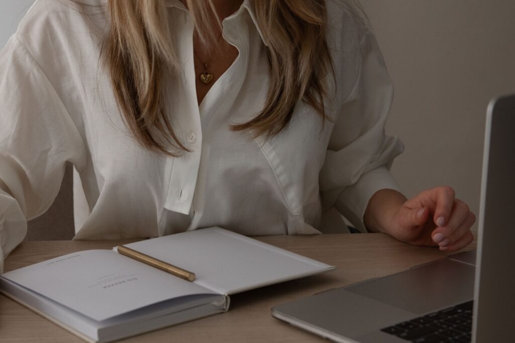 A blonde woman in a white shirt is sitting at the desk with a notepad and laptop.